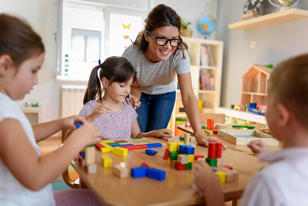 A teacher with students at a pre-school table. The children are playing with blocks.