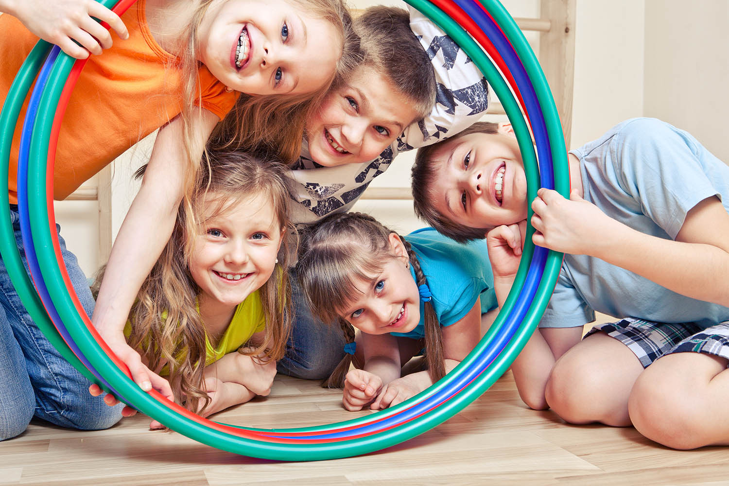Five playful kids all looking through a set of hula hoops standing upright. The kids are smiling.